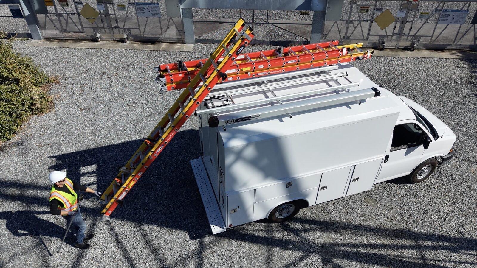 A technician in a hard hat and safety vest lowering a ladder to ground level from a white utility service van fitted with the G2000 hydraulic-assisted ladder rack